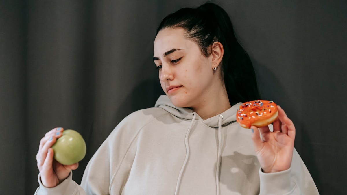 Pensive female wearing casual hoodie deciding between healthy green apple and sweet doughnut in studio